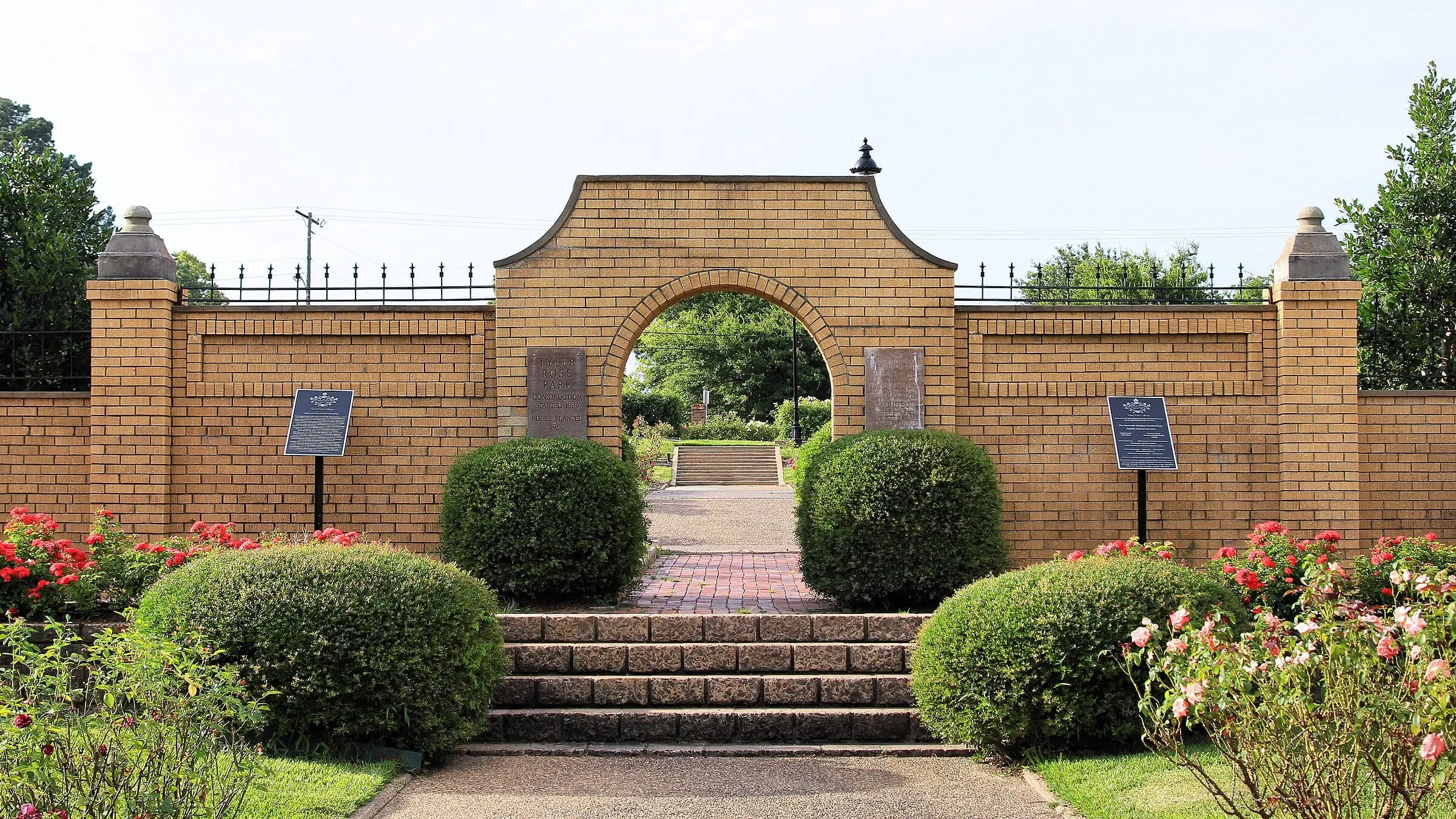 A yellow-brick wall with an arched opening at the top of a flight of brick steps; plaques on the wall and on posts, clipped shrubs, and red and pink roses in front.