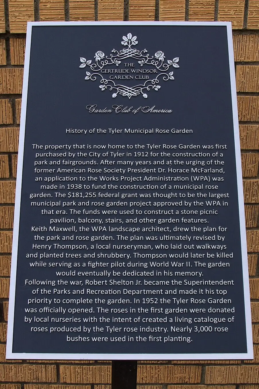 A dark plaque on a brick wall, headed History of the Tyler Municipal Rose Garden, under the emblem of the Gertrude Windsor Garden Club and the Garden Club of America, with several paragraphs of text.
