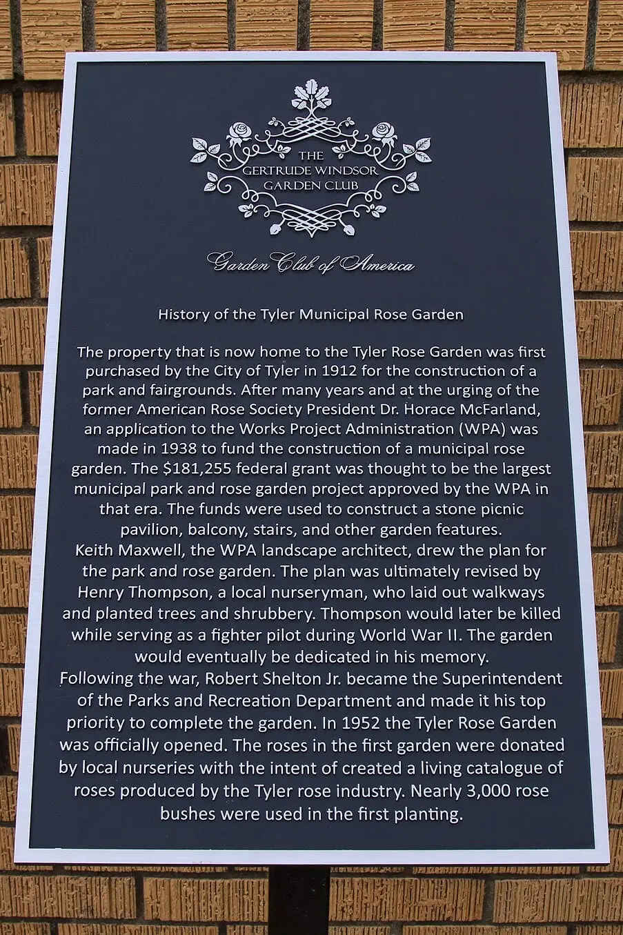 A dark plaque on a brick wall, headed History of the Tyler Municipal Rose Garden, under the emblem of the Gertrude Windsor Garden Club and the Garden Club of America, with several paragraphs of text.