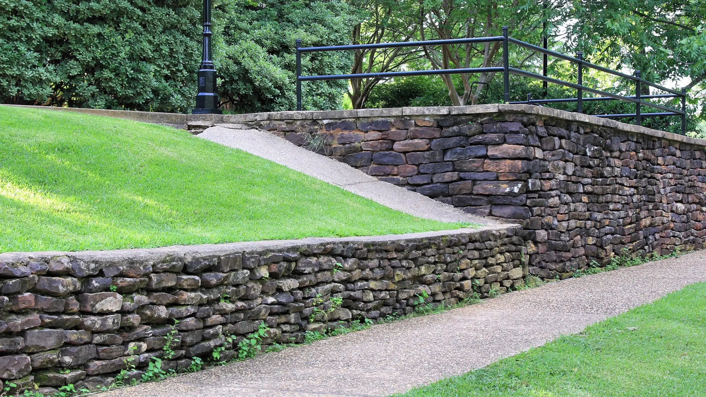 Two stone retaining walls step down a slope beside a lawn, with a walk climbing between them and an iron railing above.